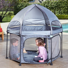 Load image into Gallery viewer, Two young children playing inside a large grey hexagonal playpen (PlayPod) with a dome canopy, set up on a patio near a pool.
